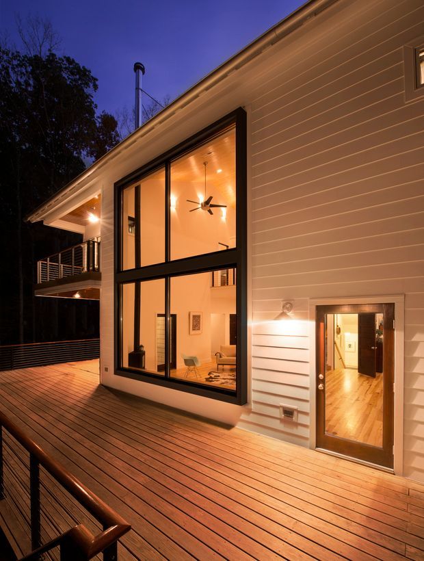 Twilight exterior view of a contemporary multi-level home from a broad timber deck. The facade is clad in horizontal light-coloured weatherboards, with expansive black-framed floor-to-ceiling glazing revealing a warmly lit interior and sculptural ceiling fan. A glazed timber entry door, balcony, chimney flue, deck railing and built-in deck seating are visible, with surrounding mature trees in the background.