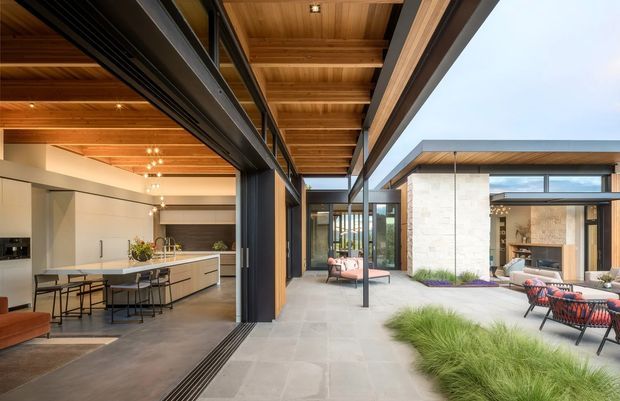 Contemporary indoor-outdoor living space organized around a landscaped courtyard. The image shows an open-plan kitchen and dining area on the left, connected through large sliding glass doors to a paved outdoor terrace and lounge on the right. Warm timber-lined ceilings, dark metal framing, pale stone or concrete paving, and broad clerestory glazing create a refined, spacious architectural composition.