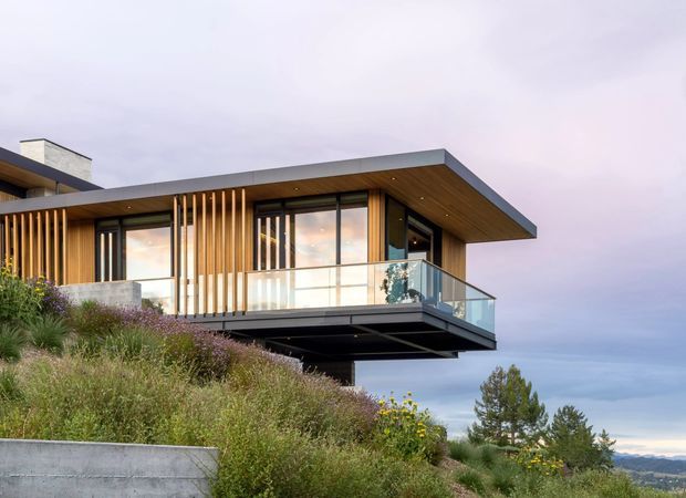 Modern cantilevered residential building on a sloping landscaped site, featuring a broad flat roof with deep overhangs, warm timber soffit and vertical timber screening, extensive glazing, and a transparent glass balcony balustrade. The elevated terrace projects over the hillside, with distant mountain scenery visible in the background.