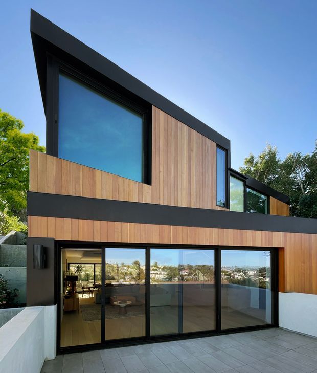 Contemporary multi-level residential facade with warm vertical timber cladding, bold black roof and horizontal trim bands, and extensive black-framed glazing. A paved terrace and low rendered walls occupy the foreground, while interior living spaces are visible through the ground-floor glass doors. The angular roofline and strong geometric composition are set against a clear sky and established vegetation.