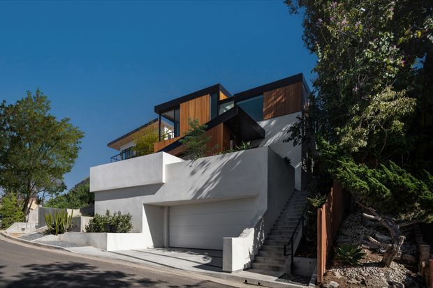 Street-level exterior view of a multi-level contemporary residence on a sloped site. The house features bold white rendered/concrete-like volumes, a prominent integrated garage, external stairway, dark structural framing, warm timber wall panels, large glazing, and substantial mature landscaping. The composition emphasizes the facade and stepped relationship to the street.