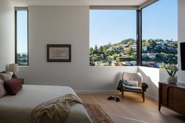 Bright contemporary bedroom with a neatly made bed, warm timber flooring, a mid-century style armchair, framed wall artwork, and a low wooden dresser. Large corner windows provide expansive views across a leafy, densely built hillside neighbourhood. The room has a restrained neutral palette and abundant natural light.