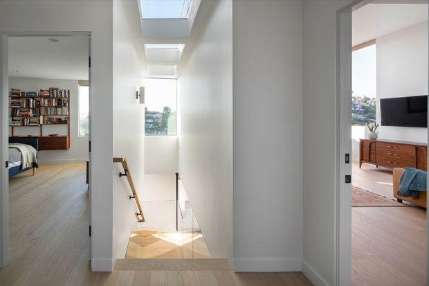 Bright contemporary residential hallway and stair landing viewed from an upper level, with a skylit stairwell descending through the centre. Open doorways reveal a bedroom with extensive built-in bookshelves on the left and a living room with a wall-mounted television and timber sideboard on the right. The space has white walls, pale timber flooring, glass stair balustrading and abundant natural light from roof skylights and a tall window.