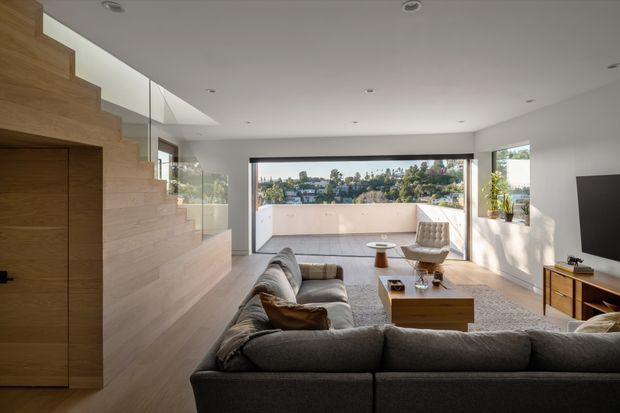Bright contemporary living room with a large grey sectional sofa, timber coffee table and media cabinetry, and a sculptural lounge chair. A timber-clad stair with integrated joinery lines the left side, while full-width glazing opens to a balcony or terrace and a leafy urban view. The room has a white ceiling with recessed downlights, pale timber flooring and strong indoor-outdoor connection.