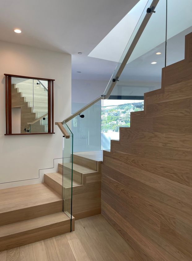 Interior view of a contemporary stairway with broad light timber treads and matching timber-clad stair walls. Clear glass balustrades with slim metal fittings and a timber handrail create an open, minimalist composition. A large wall mirror reflects the staircase, while recessed ceiling lights and a rear window provide additional brightness.