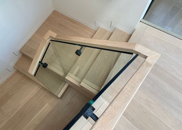 Interior view looking down a contemporary stairway with light timber treads and matching timber balustrade framing a clear glass guard. The staircase turns at a landing, with dark metal handrail or fixing details and white painted walls. Light timber flooring continues beyond the stairs.