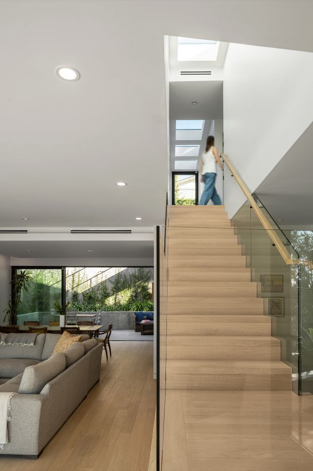 Bright contemporary interior view centred on a straight timber staircase with a glass balustrade and pale timber handrail. The stair rises through a tall void with multiple skylights and roof glazing, while an open-plan living and dining area with large windows, upholstered sofas, and timber flooring is visible to the left. White ceilings feature recessed downlights and linear air-conditioning vents.