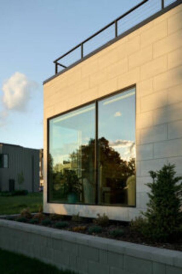 Exterior view of a contemporary flat-roof building with a pale cream rendered or masonry facade, large black-framed floor-to-ceiling glazing, and a rooftop terrace protected by a simple dark metal railing. A low retaining wall and landscaped planting sit in front of the building.