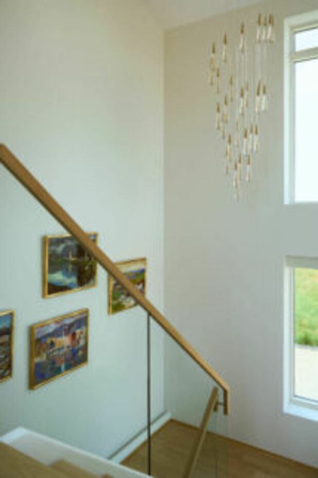 A bright, double-height stairwell with pale walls, timber stair treads and handrail, and a clear glass balustrade. Several framed artworks decorate the stair wall, while tall windows bring in abundant natural light. A delicate hanging installation adds a subtle decorative feature near the upper window.