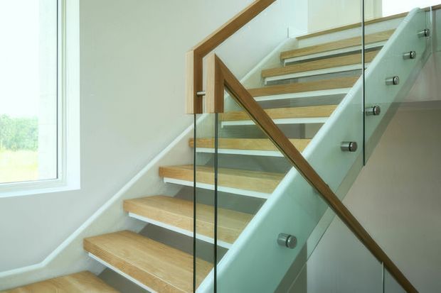 Interior view of a modern stairway with open timber treads, white side walls, clear glass balustrades secured with metal fittings, and a timber handrail. A large window provides abundant natural light and a view of greenery outside.