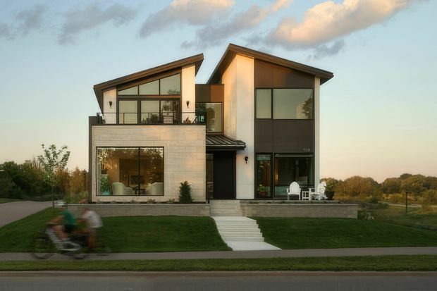 Front exterior view of a contemporary multi-storey residence with asymmetrical pitched roof forms, expansive dark-framed glazing, pale masonry cladding, and a raised entry terrace with steps. The house sits in a landscaped suburban setting beside a paved street and open greenery, photographed in warm late-day light.