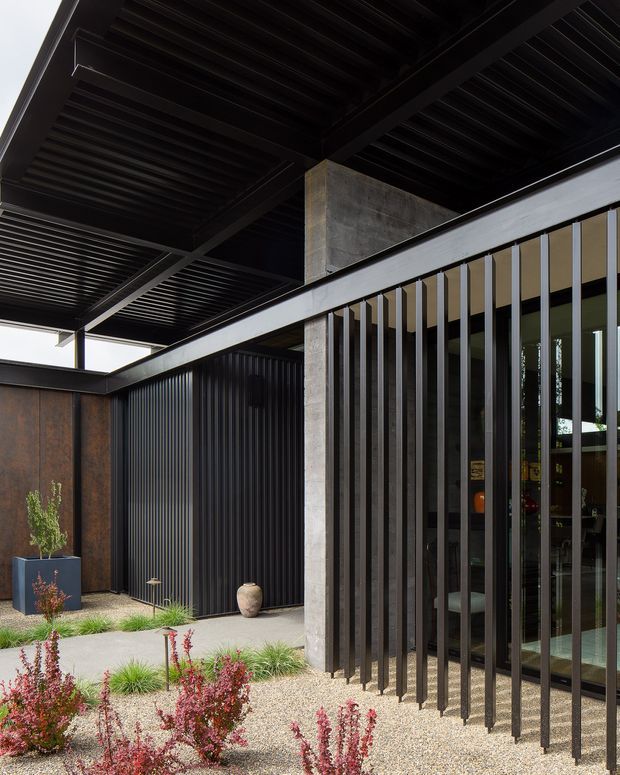 Contemporary architectural exterior showing a covered entry or courtyard framed by a bold black steel pergola and exposed concrete column. Vertical black metal fins screen full-height glazing, while dark ribbed cladding and weathered rust-toned panels create a dramatic, industrial material palette. Gravel landscaping, sculptural planting, and a paved path complete the restrained modern composition.