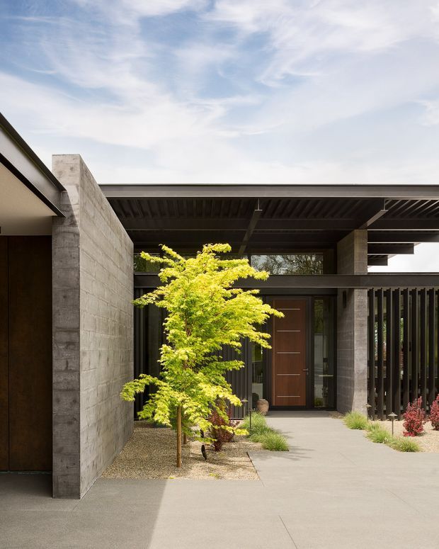 Modern single-storey residential entry courtyard viewed from outside, featuring a broad flat roof with exposed dark metal framing, raw concrete block walls and columns, dark vertical screening, and a warm timber front door. A paved concrete approach surrounds a minimalist gravel garden with a small Japanese maple and ornamental grasses. The composition is rectilinear and understated, with strong indoor-outdoor threshold design.