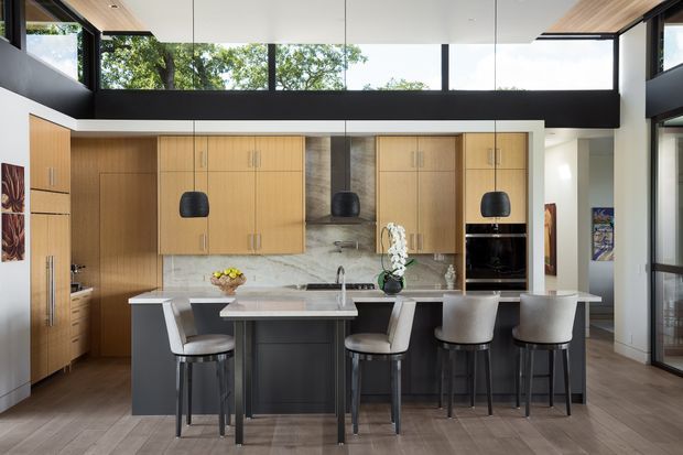 A spacious contemporary kitchen with a large central island and integrated dining-height bench. Warm timber cabinetry spans the back wall and includes tall storage and built-in appliances, while a pale marble or stone backsplash and benchtop contrast with the dark island base. Five upholstered bar stools sit around the island. Black pendant lights, dark window framing, clerestory glazing, and timber-look flooring contribute to a refined, light-filled interior.