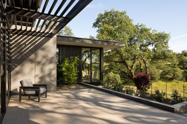 A modern outdoor terrace adjoining a glass-walled house, with a broad paved deck, dark metal pergola beams, concrete-toned walls, and a single lounge chair. The terrace overlooks a landscaped garden with mature trees and planted raised beds. Strong sunlight creates graphic shadows across the walls and paving.