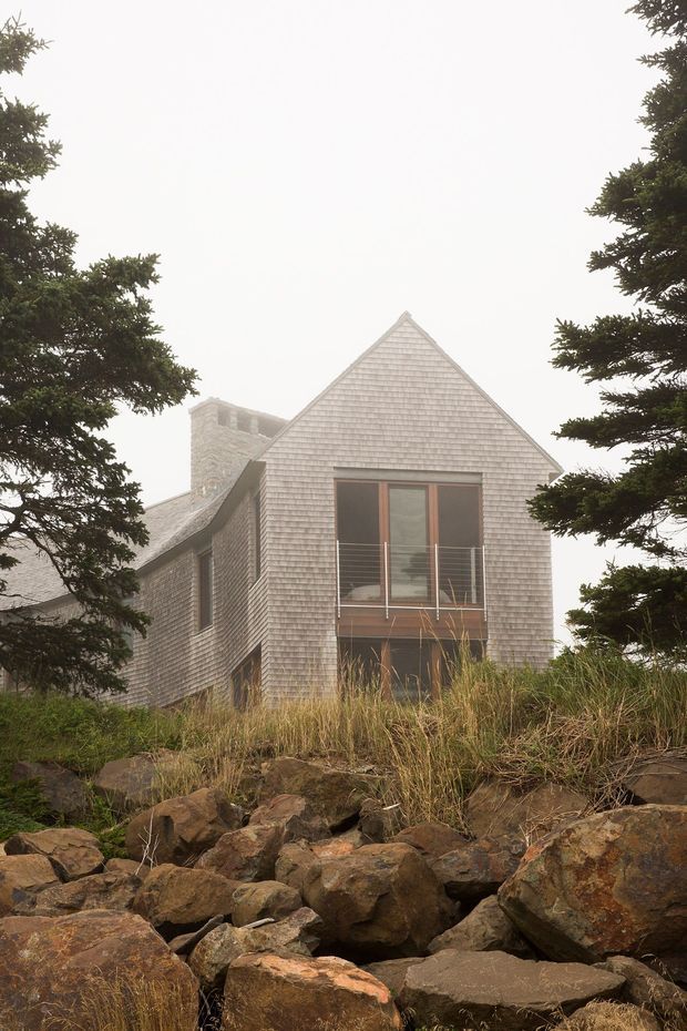 Exterior photograph of a multi-level house set among coastal grasses, rocky terrain, and evergreen trees. The building has steep gabled roof forms, shingle-clad walls and roof, a prominent central gable, dark-framed glazed doors or windows, and a masonry chimney. The overcast, misty setting gives the image a subdued natural atmosphere.