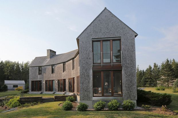 Exterior view of a substantial two-storey residence with a steep triangular gable and a long adjoining wing. The building is clad in grey shingle-like siding, with dark timber-framed glazing and a slate-like pitched roof. Landscaped lawns and garden beds surround the house, with mature trees in the background. The composition highlights the facade, gable form, and large vertical windows.