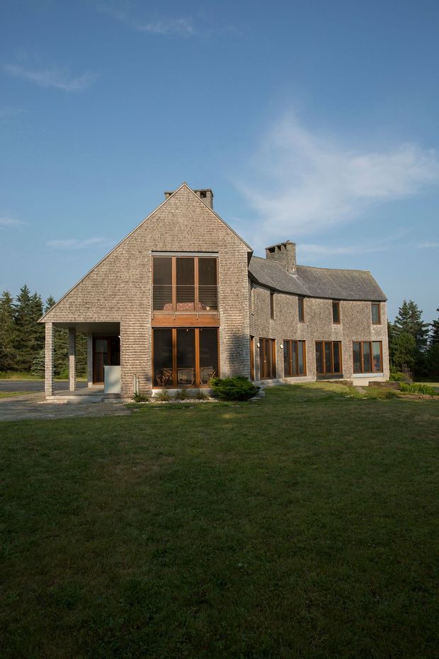 Exterior photograph of a substantial two-storey rural residence with steep gabled forms, textured shingle-clad walls and roof sections, tall timber-framed glazing, stone chimneys, and a covered entry or carport-like projection. The house sits within a broad landscaped lawn with trees at the perimeter under a clear sky.