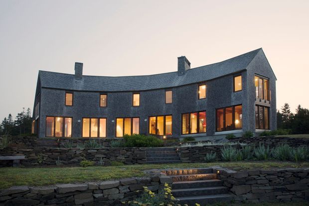 Twilight exterior view of a large curved, two-storey residence clad in dark slate-like masonry, with a steep pitched roof, multiple chimneys, extensive warm-lit windows and doors, and a projecting glazed corner bay. Terraced landscaping uses layered dry-stacked stone retaining walls and steps leading toward the house.