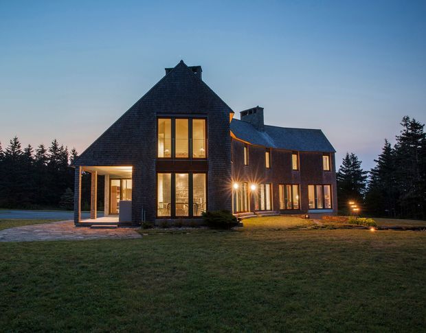 Exterior dusk view of a large contemporary house in a wooded setting. The home has steep pitched and gabled roof forms, dark textured shingle cladding, extensive warm-lit floor-to-ceiling glazing, and a projecting glazed entry or porch. A broad lawn, paved terrace, exterior lighting, and surrounding evergreen trees frame the building.