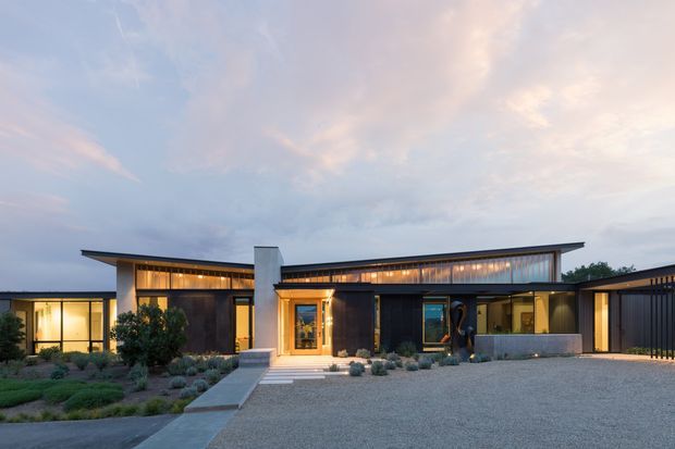 Single-storey contemporary residence photographed at dusk, with a low, asymmetrical roofline, dark cladding, warm timber-toned glazing, and broad illuminated windows. A central entry porch and concrete path face a large pale gravel forecourt, while low drought-tolerant landscaping frames the facade.