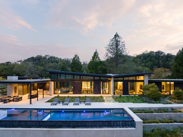 A wide dusk exterior view of a low, single-storey contemporary residence set within a wooded landscape. The home has long horizontal rooflines, extensive floor-to-ceiling glazing, dark window frames, warm illuminated interiors, and a covered outdoor living area. A rectangular swimming pool with illuminated blue water occupies the foreground, with stepped lawn and planted terraces between the house and pool.