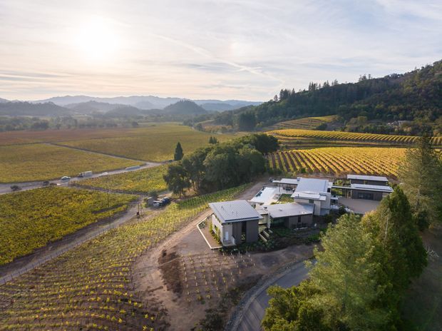 Aerial view of a modern low-rise building complex set among expansive cultivated vineyards, with patterned rows covering rolling hills and a wooded ridge beyond. The buildings have light-coloured walls, dark metal pitched roofs, and a cluster-like rural campus arrangement connected by paved access roads.