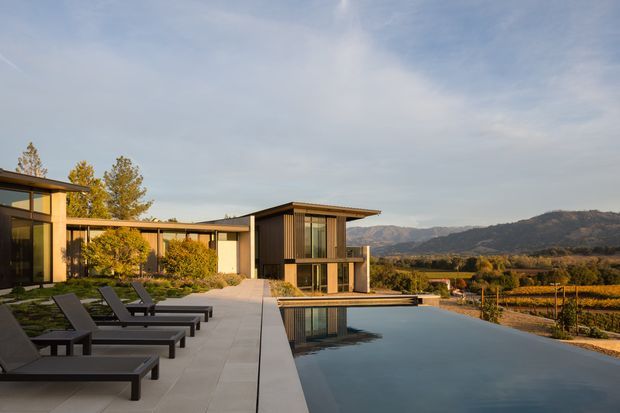 A contemporary low-profile residence set beside a long rectangular infinity pool, with a broad paved terrace and rows of dark loungers in the foreground. The house features flat roof planes, extensive glazing, warm timber cladding, and pale rendered walls. Landscaped planting and cultivated fields extend toward wooded hills and mountains in the distance, creating a refined indoor-outdoor setting.