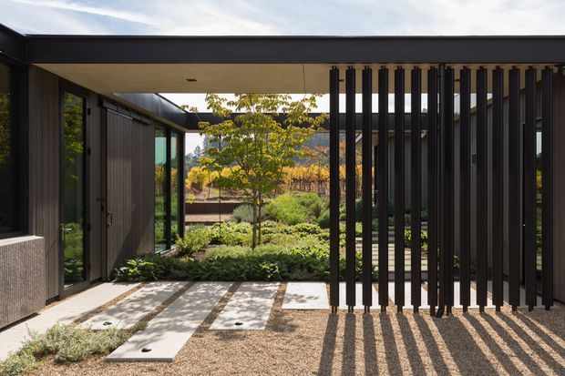 A contemporary covered outdoor passage or entry courtyard framed by dark vertical cladding, glazing, and a flat roof. A screened section of closely spaced black vertical battens casts strong linear shadows across pale concrete stepping slabs and gravel. Lush landscaped planting and a leafy tree are visible beyond the courtyard, with a dark architectural structure enclosing the space.