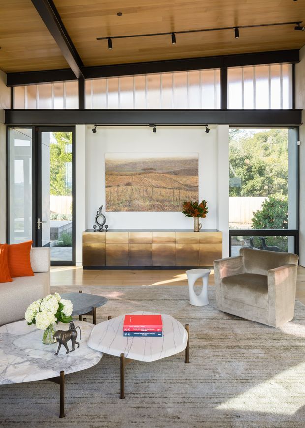 A bright, refined living room with expansive black-framed glazing, clerestory windows, exposed dark structural beams, and a warm timber-lined ceiling. The room features a large neutral rug, sculptural coffee tables, upholstered lounge seating, a bronze-toned built-in sideboard, track lighting, and a landscape artwork. Strong indoor-outdoor connection is visible through the full-height windows and glazed door.