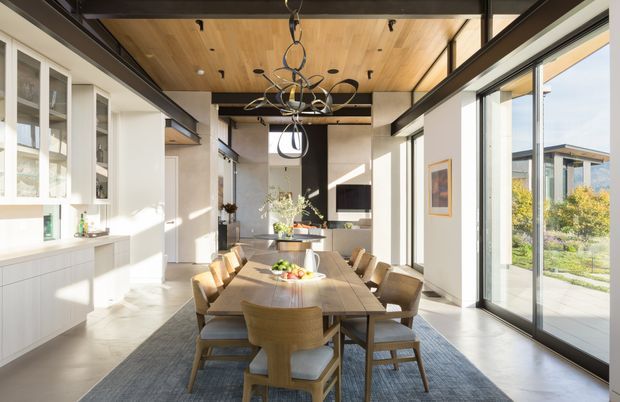 A bright, open-plan dining area with a long timber dining table and upholstered wooden chairs, adjoining a streamlined white kitchen. Floor-to-ceiling glazing opens toward a landscaped exterior, while dark metal framing, a warm timber ceiling, and a sculptural contemporary pendant light define the space.