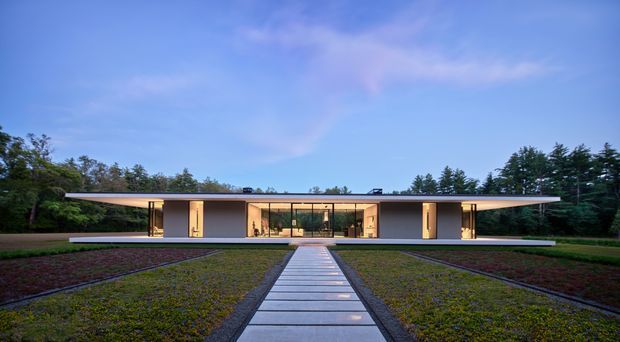 Wide frontal exterior view of a low, symmetrical contemporary house set within landscaped grounds. The building has a long flat roof with broad overhangs, dark horizontal roof and fascia elements, light rendered walls, extensive floor-to-ceiling glazing at the central entrance and living areas, and a straight paved path leading to the entrance. Dense trees frame the property under a blue evening sky.