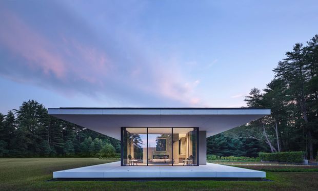 A minimalist single-storey pavilion-like house set within a landscaped, wooded site. The structure has a broad flat roof with deep cantilevered overhangs, full-height glazed walls, and a clean white or pale-grey platform terrace. Warm interior lighting reveals a sparsely furnished living space, while mature trees and lawn surround the building at dusk.