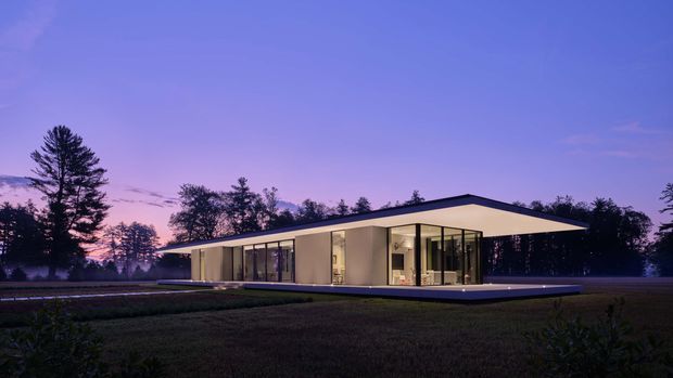 A low, minimalist single-storey modern house set in a landscaped, wooded setting at dusk. The building has a broad shallow roof, crisp white or pale rendered walls, extensive floor-to-ceiling glazing, and a projecting terrace or plinth. Warm interior lighting contrasts with the cool purple-blue evening sky and dark surrounding trees.