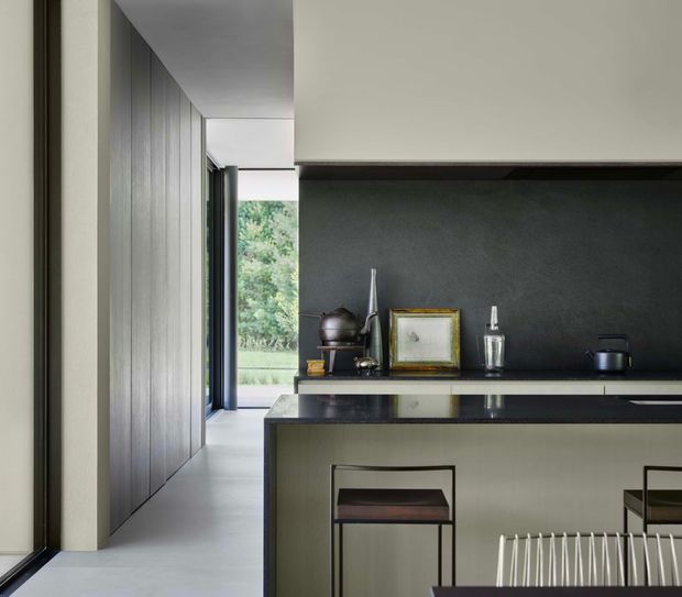 A refined contemporary kitchen viewed from the adjacent circulation space. The room features a dark charcoal stone-look splashback, glossy dark benchtop, pale textured fixed cabinetry, and a long island with minimalist black-framed stools. A tall timber-panelled wall and floor-to-ceiling glazing lead toward greenery outside, while restrained artwork and objects sit along the rear counter.