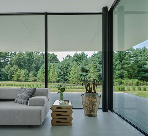 A contemporary living room with expansive floor-to-ceiling glazing overlooking a landscaped garden. The space features a pale upholstered sofa, a sculptural stacked side table, a large ceramic vessel, and dark slender window frames and structural posts. A broad paved terrace is visible outside.