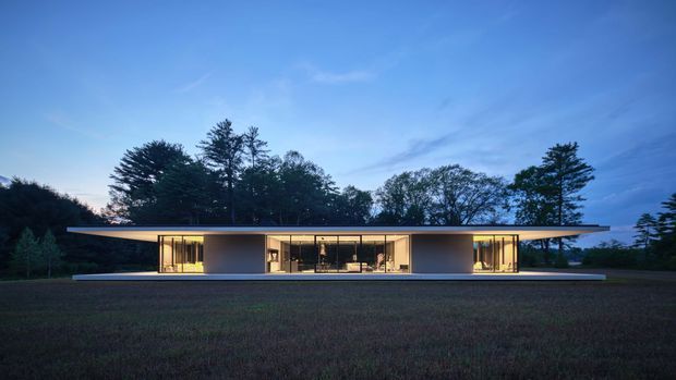 Wide dusk exterior view of a long, low-slung modern house set within a wooded landscape. The residence has a dramatic thin overhanging flat roof, extensive floor-to-ceiling glazed openings, warm interior lighting, and a minimalist horizontal composition with a broad open lawn in front.