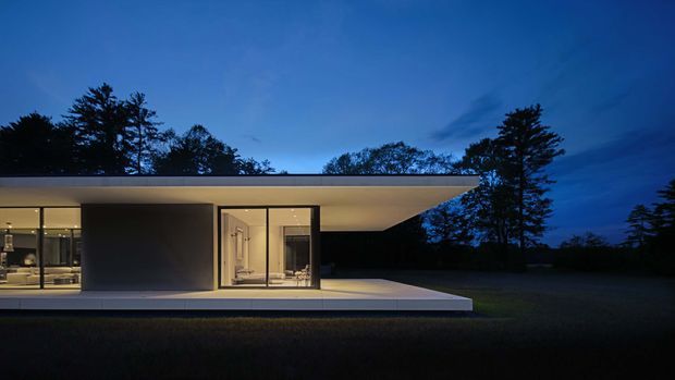 A low, minimalist modern residence photographed at dusk, with a broad flat roof projecting over a glazed living area and recessed dark wall volumes. Floor-to-ceiling glazing reveals a warmly lit interior, while a pale paved terrace extends across the foreground and mature trees form a dark backdrop.