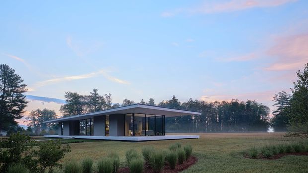 A low, single-storey contemporary pavilion-style house set in an open landscaped rural or suburban site. The building has a broad flat roof with deep overhangs, extensive floor-to-ceiling glazing, dark slender window frames, and a restrained grey facade. Ornamental grasses and low planting surround a broad paved terrace, with trees and soft evening light in the background.