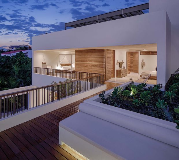 Twilight exterior view of a modern multi-level residence with a crisp white rendered facade, broad open-plan glazing, warm timber-clad interior surfaces, and extensive timber decking. Slim metal balustrades line the elevated terrace and walkway, while integrated planters and lush planting soften the architectural forms. The image also reveals a warmly lit interior living area through the open facade.