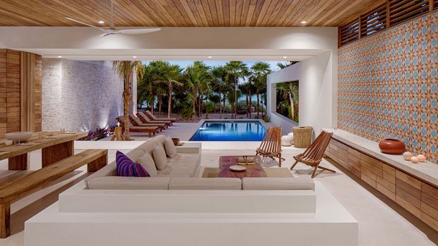 A refined open-plan indoor-outdoor living space overlooking a rectangular swimming pool and tropical garden. The room features a large cream sectional sofa, timber dining table and benches, woven lounge chairs, a timber slatted ceiling, white structural walls, built-in timber cabinetry, and a striking blue-and-terracotta patterned tiled feature wall. The architecture creates a strong framed connection between the living room and pool terrace, with a bright, warm, resort-like atmosphere.