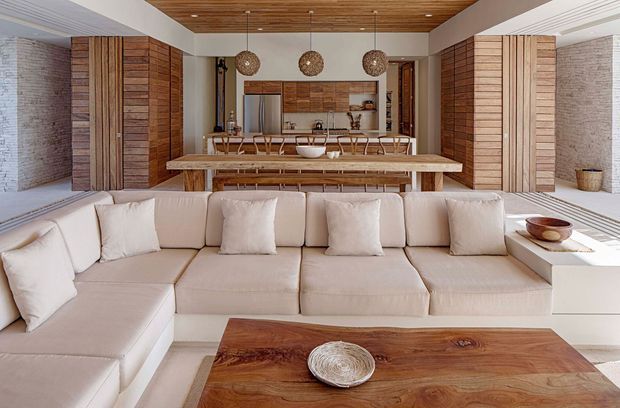 Open-plan living and dining area with a large cream sectional sofa in the foreground, a substantial natural timber coffee table, a long dining table with woven chairs, and a compact kitchen beyond. The space features extensive warm timber wall panelling and cabinetry, a white painted brick feature wall, a timber slatted ceiling, and three woven pendant lights. The overall design is warm, textural, and contemporary with strong natural-material detailing.