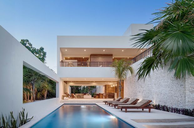 Contemporary multi-level residence viewed from a pool courtyard, featuring clean white geometric volumes, open-plan glazed living areas, a covered upper terrace with timber screening, and a long rectangular swimming pool bordered by timber sun loungers. A textured white stone wall and tropical planting frame the courtyard, creating a refined resort-like outdoor setting.