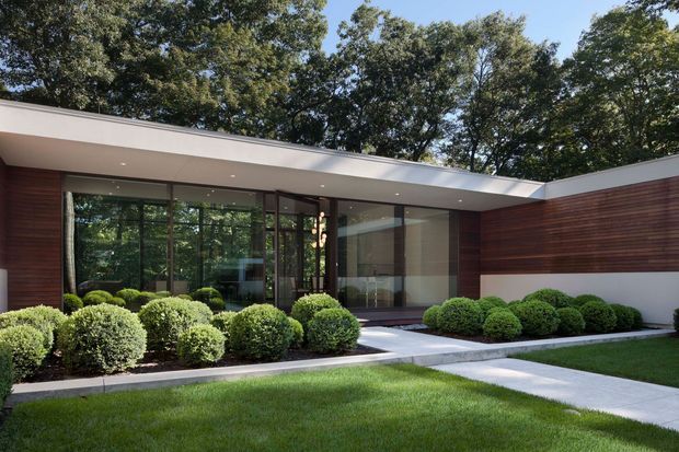 A low-slung modern single-storey residence viewed from the landscaped front garden. The house has a broad flat roof with recessed downlights, extensive floor-to-ceiling glazing in dark frames, and horizontal dark timber wall cladding contrasted with smooth white rendered walls. Rounded clipped shrubs line the facade, with a paved walkway crossing a manicured lawn.