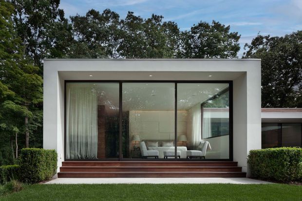 Exterior view of a refined modern single-storey residence set within landscaped greenery. The prominent white rectangular pavilion has a flat roof, expansive dark-framed floor-to-ceiling glazing, broad timber entry steps, and a bright living room visible through the windows. A lower adjoining wing extends to the right, with clipped hedges and lawn in the foreground.