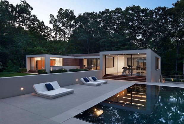 A modern, single-storey flat-roof residence set within a wooded landscape. The home features clean rectilinear volumes, extensive floor-to-ceiling glazing, warm timber wall cladding, and a broad concrete terrace surrounding a reflective swimming pool. Minimal outdoor loungers, integrated terrace lighting, and the evening setting create a refined, tranquil atmosphere.