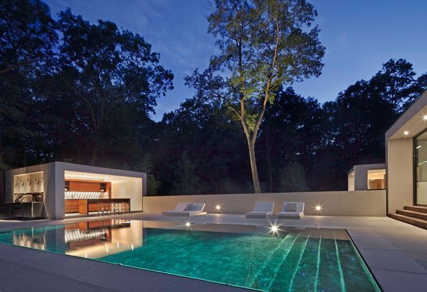 Dusk view of a luxurious contemporary pool terrace surrounded by mature trees. A large illuminated swimming pool dominates the foreground, with sculptural loungers and low rendered walls around it. Open-plan pavilion-like structures with strong rectangular forms, warm timber-lined interiors, integrated lighting and broad glazing frame the pool, while steps lead into a bright modern building at right.