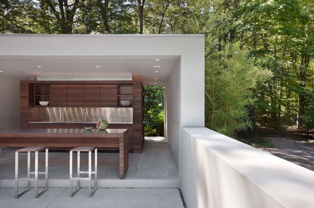 A contemporary outdoor kitchen pavilion set within a wooded landscape. The open, white-rendered structure contains dark timber cabinetry, a stainless-steel backsplash and benchtop, a large island with two metal stools, and pale concrete flooring. A low white parapet frames the right side, with recessed ceiling lights and a leafy forest visible beyond.