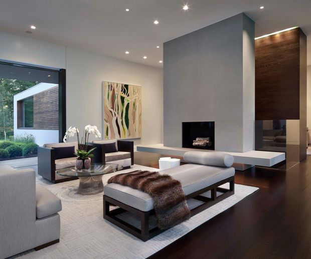 Contemporary living room with a large pale grey central fireplace wall, linear built-in media or display cabinetry, dark timber flooring, and recessed ceiling lighting. The room is furnished with a large upholstered ottoman, lounge seating, glass coffee table, abstract artwork, and a neutral textured rug. Full-height glazing at left looks onto a landscaped exterior and a modern timber-clad structure.
