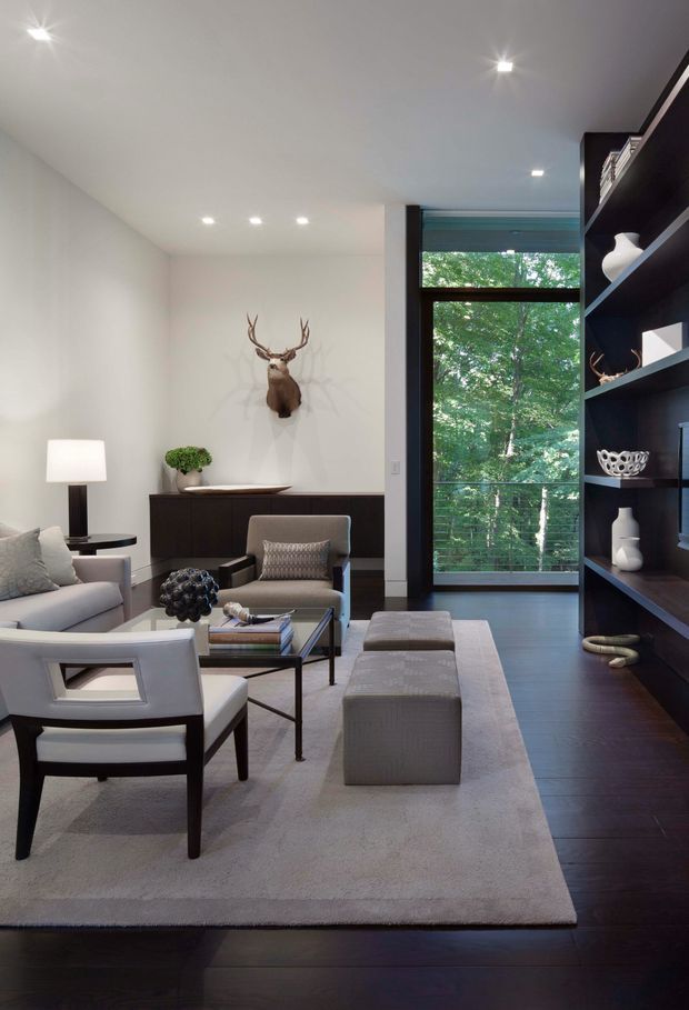 A refined contemporary living room with a neutral palette, dark timber flooring, pale upholstered seating, a large light rug, and a built-in dark shelving wall displaying decorative objects. A black-framed floor-to-ceiling window provides views of trees, while recessed ceiling lights and a wall-mounted deer sculpture add visual interest.